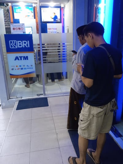 The photo shows an ATM in Sarimulyo, Indonesia, where two men are looking at their cell phones. The ATM is for BRI bank. The man in the front wears a dark blue shirt and khaki shorts with dark sandals. The man behind him wears a light blue shirt, brown sarong, and a dark hat. The floor is tiled, and there is a blue door mat. The door to the ATM is glass. The signage on the ATM includes the bank's logo and accepted payment methods. The time of day appears to be late afternoon or early evening.