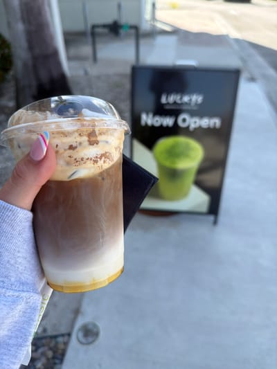 A person's hand, with manicured nails featuring pink, white, and blue stripes, holds a clear plastic cup filled with a layered iced coffee. The drink has distinct layers of white at the bottom, a caramel-colored liquid in the middle, and a foamy, creamy topping with ice cubes. The cup is partly obscured by a dark object. In the background, a black A-frame sign with "Lucky's" and "Now Open" in white lettering is visible. To the right of the sign, a blurry green beverage in a cup is also visible. The scene appears to be outdoors, on a sidewalk with a grey concrete surface, suggesting a commercial area. The lighting indicates it is daytime. This scene is likely located in North Miami Beach, United States of America, given the context of the establishment advertised on the sign.