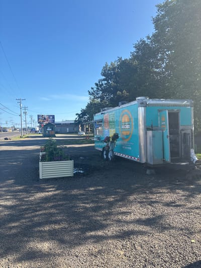 A light blue food truck named "Recess" is parked on gravel next to a street, with a tree behind it. The truck has a menu displayed on the side and is equipped with a small wheeled cart adorned with flowers. A white planter box with flowers sits nearby. Power lines run along the street, and buildings are visible in the distance. The sky is clear blue.