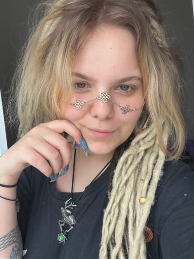 The media file shows a close-up, low-angle, sideways shot of a person with blonde dreadlocks, possibly a young woman, looking towards the camera. She has fair skin with freckles, blue eyes, and light pink lips. A delicate silver chain with Celtic knot-like charms is worn across her forehead and cheek. Her fingernails are painted a dark blue-green with a marbled effect. She is wearing a black t-shirt, visible on which is a silver pendant of a deer head with antlers, holding a green orb in its mouth. The background is dark and out of focus, suggesting an indoor setting. The lighting is soft, and the overall mood appears casual and perhaps a bit mystical due to the jewelry and pendant. There is no visible text in the media file. The location context provided is Vostochny District, Russia.