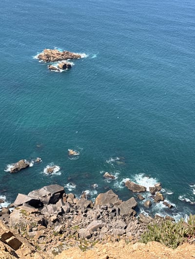 The image is a high-angle shot of the Atlantic Ocean off the coast of Colares, Portugal, likely taken from Cabo da Roca, the westernmost point of mainland Europe. The scene is dominated by the vast expanse of deep blue-green water, with gentle ripples on its surface. Several rock formations are visible, ranging from small, wave-battered outcrops closer to the shore to larger, more substantial islands further out at sea. The foreground is occupied by a steep, rocky cliff face, composed of various shades of brown and gray stones. Sparse, green, succulent-like vegetation grows in patches among the rocks on the cliff. The overall impression is one of a rugged, natural coastal landscape under a bright, clear sky. There are no people or visible signs of human activity in the image, other than the implied vantage point from a cliffside observation area.