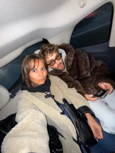 Two young adults, a man and a woman, are captured in a rotated, eye-level indoor shot within what appears to be a car, specifically in the Saint-Laurent-du-Var, France area. The man, on the left, has curly light brown hair and a beard, wearing sunglasses, a grey hoodie, and a dark brown furry jacket. He is holding a smartphone in his right hand, displaying what looks like a screen with multiple small images. The woman, positioned closer to the foreground, has dark brown hair with bangs, wearing a black hooded top and a light beige fluffy jacket. Both individuals are looking directly at the camera with neutral to slightly serious expressions, with the woman's eyes wide open. A black handbag or purse is visible near the woman. The setting suggests they are in the back seat of a vehicle, possibly at night or late evening, as indicated by the dark exterior visible through the car window, where a red light from another vehicle's taillights can be faintly seen. No discernible text is visible in the image.