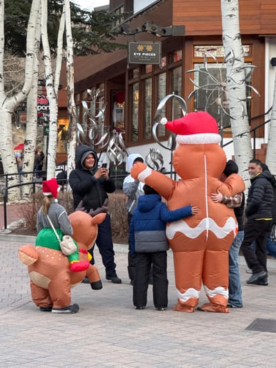 A group of people wear inflatable costumes, including a gingerbread man with a Santa hat and a reindeer, walking along a brick sidewalk next to aspen trees. A man in a hooded jacket smiles at the camera. A store called "Eye Pieces" is visible in the background. People in casual attire walk by in the distance.
