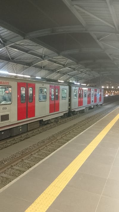 A subway train with red doors and silver cars sits at a station. Passengers are visible inside the train. The station has a high, arched metal roof with lights. A yellow tactile strip runs along the gray platform edge. Train tracks with gravel fill the space between the train and platform. Outside the station, street lights are visible in the distance.