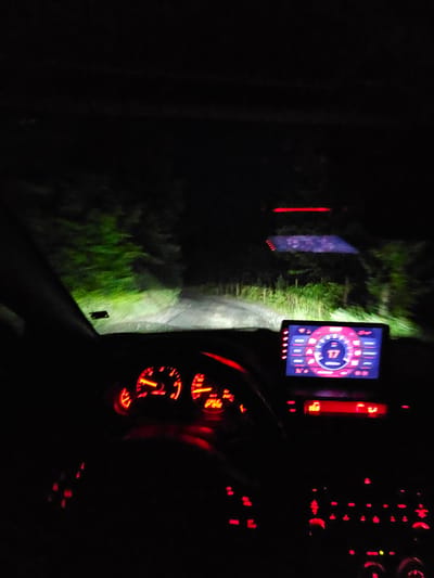 The image appears to be taken from inside a vehicle at night. The dashboard with red illuminated gauges is visible, as well as a digital display showing numbers and a circular graphic. Through the windshield, a road is illuminated by headlights, leading into a dark, wooded area. There are no people visible in the image. The location is Erma Reka, Bulgaria. The image suggests the vehicle is driving on a road surrounded by foliage at night.
