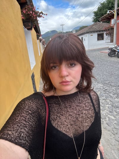 A young woman with brown hair and bangs poses for a selfie outdoors. She wears a black, sheer top and a delicate necklace. The background shows a cobblestone street, a yellow building with white accents and potted plants, and a distant mountain under a partly cloudy sky. A motorcycle is parked on the street. The image is taken from a slightly high angle and has been rotated.