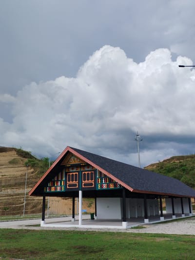 The image shows an outdoor scene with a building featuring traditional architecture in Keureuweungkrueng, Indonesia. The building has a dark-colored, pitched roof and is supported by dark-colored pillars. The front facade of the building is decorated with colorful geometric patterns and what appear to be windows. To the left of the building, a terraced hillside is visible, with some vegetation on its slopes. In the background, there are more hills and a large expanse of cloudy sky. A street lamp is visible in the upper right portion of the image, and another tall pole-like structure with lights is present further in the background. The ground in front of the building is covered with grass and a paved area. The overall impression is of a well-maintained public space with a focus on cultural architectural elements.