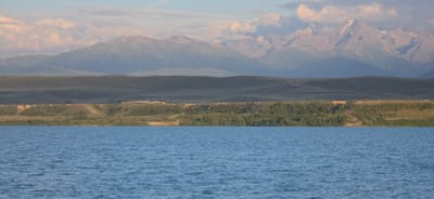 The image captures a serene natural landscape with a body of water in the foreground and mountains in the background, under a soft, cloudy sky. The water is a vibrant blue, rippling gently, suggesting a peaceful atmosphere. Beyond the water, a green, tree-covered slope rises, transitioning into rolling, grassy hills that recede into the distance. These hills are muted in color, indicating a considerable distance. The furthest element of the landscape is a majestic mountain range, its peaks sharp and rugged, with some snow visible on the higher altitudes. The mountains are bathed in the warm, soft light of either sunrise or sunset, casting a gentle glow and creating a hazy, atmospheric effect. A few clouds are present in the sky, tinged with pink and orange from the ambient light. The overall scene evokes a sense of tranquility and vastness, characteristic of remote natural environments. The location is identified as Pristan'-Przheval'sk, Kyrgyzstan, a region known for its stunning natural beauty, including large bodies of water and imposing mountain ranges. No people or man-made structures are prominently visible, emphasizing the natural focus of the image.