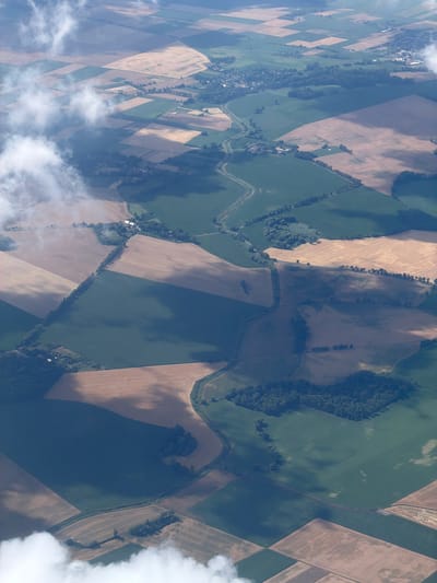 An aerial view captures a patchwork of agricultural fields in various shades of green and brown, dissected by roads and small bodies of water. Clouds drift across the landscape, casting shadows that create patterns of light and dark. The image displays a rural environment with evidence of human activity in the form of cultivated land and scattered buildings. The precise location of Raczkowa, Poland, is not discernable from the visual information presented in the image. There are no people or specific subjects depicted, only the vast expanse of the cultivated land.