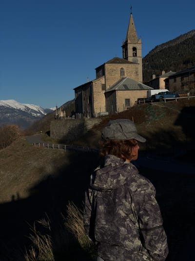 A person wearing a cap and a patterned jacket is looking at the Saint-André church, located in Saint-André, France. The church has a tall spire and a stone exterior. Mountains are visible in the background. A blue and white vehicle is parked near the church. A small road is adjacent to the church, and a grass-covered slope is in the foreground.