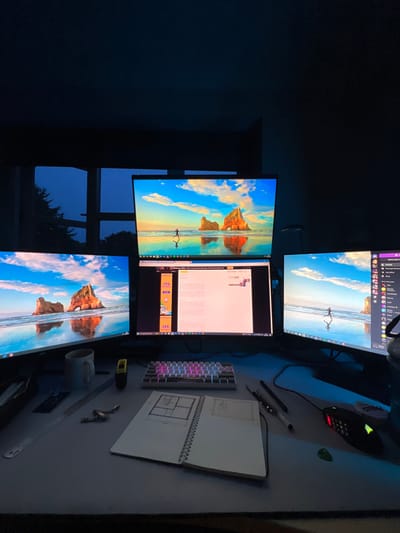 This is a forensic description of an indoor computer workstation setup located in Hornsea, United Kingdom, captured during evening or night hours.
The scene is dominated by a desk featuring a multi-monitor computer configuration. Three primary monitors are active: a large central monitor positioned above a smaller central monitor, and a monitor to the left and right. The top central monitor and the left and right monitors display an identical bright desktop wallpaper featuring a sunny beach scene with distinctive rock formations in the water and a solitary figure running near the shoreline, reflected on the wet sand. The lower central monitor displays a web browser window, showing text content and advertisements, including a prominent banner for "75% annual membership of Chinese Butter Tea." The rightmost monitor displays the Discord application interface.
On the desk surface, directly in front of the monitors, a compact mechanical keyboard with illuminated RGB keys is centrally positioned. To its right, a black gaming mouse with red lighting accents rests on a dark mouse pad. To the left of the keyboard, a light-colored mug is visible. Further to the left, a silver ruler and a small yellow object resembling a measuring tape are present. In the foreground, an open spiral-bound notebook displays hand-drawn floor plans or architectural diagrams on its left page. Several pens and pencils are scattered near the notebook and mouse.
The ambient lighting is dim, indicating low natural light, suggesting the time is either late evening or night. A window is visible in the upper background, revealing a dark blue sky and the silhouettes of coniferous trees, confirming the external environment is dark. No individuals are visible in the image. The scene depicts an active, organized workspace.