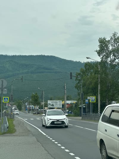 The image depicts a cloudy day in Slyudyanka, Russia, looking down a paved road that leads towards a mountainous, forested landscape. A white sedan with a license plate reading "T300XH 38" is driving in the right lane. A white minivan is parked on the right side of the road. On the left side of the road, there is a pedestrian crossing sign and a guardrail. In the background, there are several streetlights and buildings. A billboard advertising construction materials and delivery services is visible on the right side of the road. The sky is overcast with visible clouds.