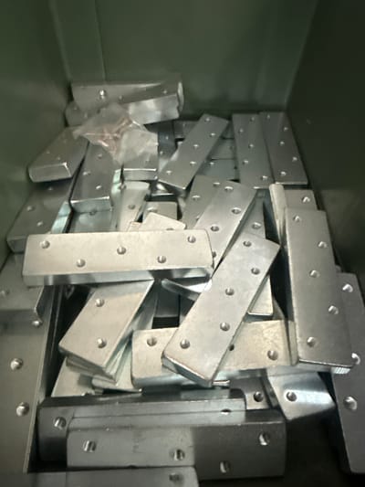 The image depicts a close-up, overhead view of a green bin filled with numerous metal brackets. The brackets are rectangular with rounded corners and have multiple circular holes, some of which appear to be threaded. They are stacked and arranged haphazardly within the bin, creating a textured surface. A small, clear plastic bag containing screws is visible on top of some of the brackets. The lighting suggests an indoor environment, possibly a workshop or storage area. There is no indication of the specific location within Fontevivo, Italy.