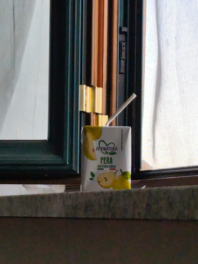 A carton of pear juice sits on a window ledge. The carton is white with an image of two pears and the text "AMONATURA PERA solo frutta italiana". A straw extends from the top of the carton. The background shows a window with vertical blinds and sheer curtains. The setting appears to be indoors, near a window. There is no indication of a specific time of day or weather conditions.