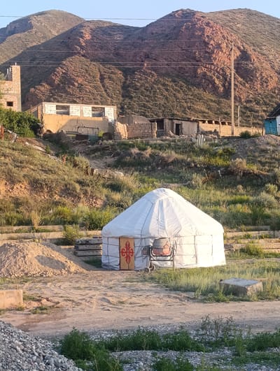 A traditional white yurt stands in the foreground of this outdoor scene, set against a backdrop of hills and undeveloped buildings. The yurt is cylindrical with a conical roof, adorned with a decorative panel featuring a red and yellow symbol. A small, orange cement mixer with wheels is parked to the left of the yurt.

Behind the yurt, the landscape slopes upward, covered in dry grass and scattered patches of greenery. Several rudimentary structures, possibly under construction or in disrepair, are visible on the hillside. These buildings are made of brick and adobe, with some lacking roofs or windows. A tall brick tower stands to the far left, partially obscured by vegetation.

In the distance, rugged, brown hills rise dramatically, their slopes etched with ravines and covered in sparse vegetation. The sky above is a clear, pale blue, suggesting fair weather. The lighting indicates it is either early morning or late afternoon, with long shadows cast by the yurt and the surrounding elements. The scene appears to be a rural or semi-developed area in Naryn City, Kyrgyzstan, characterized by its natural landscape and simple human habitation. There is no visible text in the image.
