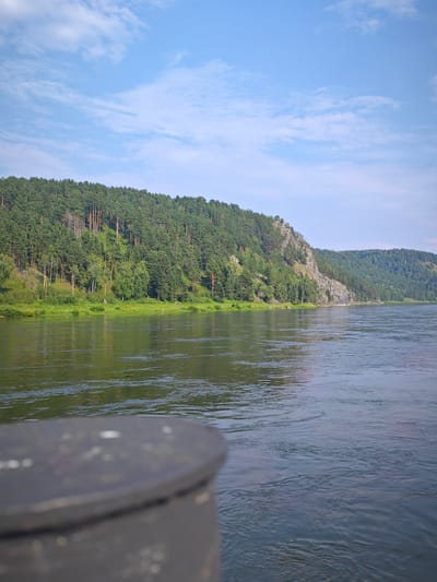 The image shows a wide river with a forested mountain range on the opposite bank, under a partly cloudy blue sky. A dark, rounded object, possibly a bollard or a part of a boat, is in the foreground, slightly out of focus. The river reflects the green of the trees and the blue of the sky. The mountain is covered with a dense pine forest, with some rocky outcrops visible. The shoreline at the base of the mountain is lined with green grass and smaller trees. The scene suggests a natural, serene environment. This appears to be a view of the Yenisei River near Beregovaya-Podemnaya, Russia, known for its scenic beauty and surrounding natural landscapes.