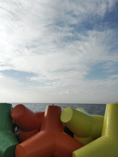 The image shows a cluster of vibrantly colored tetrapods on a coastline. The tetrapods are concrete structures designed to prevent coastal erosion. In this image, they are painted in green, orange, and yellow. They are arranged in a dense formation, with some partially submerged in the water. The ocean stretches out beyond the tetrapods, meeting a pale blue sky filled with wispy white clouds. The water appears calm with small waves. The overall scene is one of a coastal defense system situated by the sea under a partly cloudy sky. The location is identified as Phú Thủy, Vietnam. There are no people or animals visible in the image.