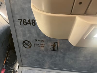 This image displays the interior of a public transportation vehicle, located within the city of Edison, United States. The scene is dominated by a textured blue-gray wall panel, marked with the black numbers '7648'. Below the numbers, three distinct black-on-white notices are present: a 'no smoking' icon (a cigarette within a circle crossed by a diagonal line), the capitalized text 'PLEASE KEEP FEET OFF SEATS.', and a 'litter' icon (a stylized human figure dropping an item into a trash receptacle within a square border). Above this panel, a curved, off-white overhead structure, possibly part of a luggage rack or ceiling, occupies the upper right portion of the frame, showing a bright, rectangular light fixture. In the lower-left corner, the lower torsos and legs of two individuals are partially visible. One person is dressed in blue jeans and a dark top or backpack, standing adjacent to the panel. Behind them, another person is partially obscured, wearing patterned clothing. No active interactions or specific actions are evident.