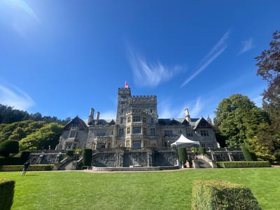 This is a full, outdoor, eye-level, wide-angle shot of a large stone castle under a bright blue sky with scattered wispy clouds. The castle, located at the Butchart Gardens in Brentwood Bay, British Columbia, Canada (near the city of Langford), features a tall central tower topped with a Canadian flag. A white tent is set up on the right side of the castle's terrace. The castle is surrounded by meticulously maintained green lawns, manicured hedges, and a stone retaining wall. Several people are visible on the castle terrace and lawn, and one person is running on the lawn in the foreground. The background consists of lush green trees and vegetation on a hillside.