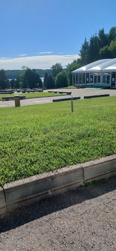 This is a vertical, eye-level, outdoor medium shot of a paved path next to a grassy area. A large white tent structure is visible in the background, with a row of trees and a hazy sky beyond. The tent has several large glass doors with white frames. The grassy area is well-maintained and features several dark, cylindrical objects positioned in a row along the path. A trash receptacle is also visible in the grass. The scene appears to be set during the day, under a clear, blue sky with some wispy clouds. The lighting suggests it is mid-day or early afternoon, and the weather appears to be fair. The foreground is dominated by the rough texture of an asphalt road and a concrete curb, with small weeds growing in the cracks of the curb. The overall impression is a peaceful, outdoor setting, possibly a park or a venue. No people are visible in the image. No text is legible in the image.