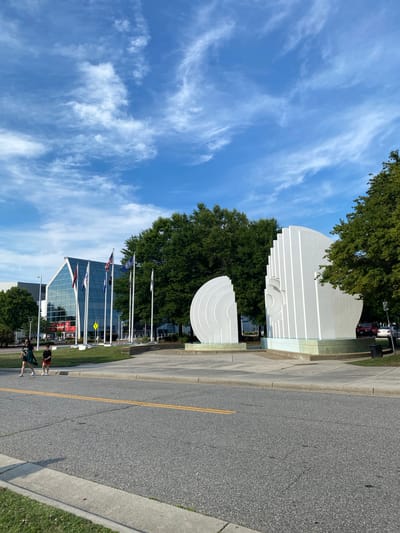 This image captures a sunny day scene in Virginia Beach, USA, featuring a large, modern building with a glass facade, several flags, and two large white sculptures. In the foreground, two people are walking across a street. The building, which appears to be a convention center or arena, has a pointed, angled roofline and displays a large advertisement on its exterior. In front of the building, a series of flags fly on tall poles, including the American flag and what appear to be state or organizational flags. The sculptures are abstract and white, with one resembling a half-circle and the other a stylized human face with vertical lines. Lush green trees form a backdrop for the scene. The sky is blue with scattered white clouds. The street in the foreground is paved with asphalt and marked with yellow lines.