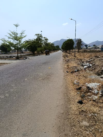 The scene, located in Rafin Pa, Nigeria, depicts a straight, two-lane asphalt road extending longitudinally into the distance under a clear, bright sky. On the left side, a strip of ground contains several trees with green foliage, some exhibiting distinctive V-shaped branching patterns. Further left, beyond the trees, indistinct low-rise structures are visible. A yellow and orange motorized tricycle is present in the left lane, positioned in the mid-distance, facing away from the viewer.
The right side of the road features an unpaved shoulder covered in dry, light brown vegetation and substantial scattered litter, including white plastic material and darker debris. A tall, silver street light pole with an active illuminated fixture stands upright on the right shoulder, approximately mid-frame. Overhead power lines are visible in the upper right quadrant of the image. One or two indistinct human figures are discernible in the far distance on the right side of the road, appearing to walk.
In the background, a range of large, rocky hills or mountains rises under a light blue sky with minimal cloud cover. The overall environment appears dry and sunlit.