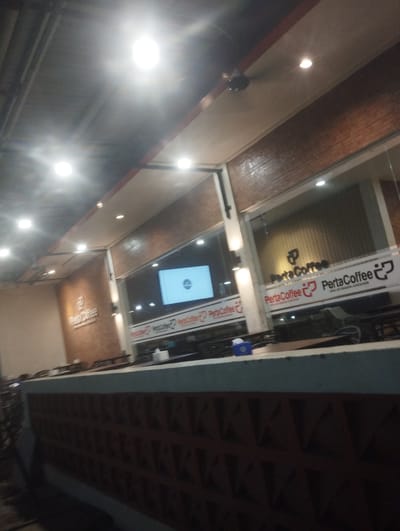 The image shows the exterior of a PertaCoffee shop. The shop has a brick facade and large glass windows. Inside, a screen displays the PertaCoffee logo. The shop is well-lit with ceiling lights. A counter is visible in the foreground. The location is Meunasahceubrek, Indonesia.