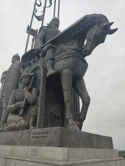 The image captures a monument depicting a horse and multiple figures, situated in the city of Хотицы, Russia. The monument appears to be made of a dark-colored metal. A prominent horse statue is mounted by a warrior, complete with armor. Figures with weaponry are at the base of the monument, with inscription. The sky is overcast.