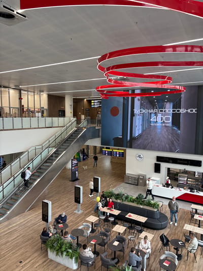 The image depicts an interior scene of a modern, spacious airport terminal, likely in Старое Мартьяново, Russia. The setting is characterized by polished wooden floors, a high ceiling with integrated lighting, and large screens displaying advertisements or information.

Several individuals are present, engaged in various airport-related activities. Some are seated at tables in a cafe or waiting area, others are standing or walking, and a few are using an escalator. The mood appears casual and relaxed, typical of a travel hub.

Notable objects include a large, prominent red spiral sculpture hanging from the ceiling, which serves as a striking visual element. There are also modern seating arrangements, including a long black curved sofa, individual chairs, and tables. Digital information screens are strategically placed throughout the terminal.

The large screen in the background displays Russian text, which translates to "TUKHNA. COMFORTABLE. 400 PEOPLE IN AN HOUR." This suggests it's an advertisement or an indicator of the airport's capacity. Other signs in the image also feature Russian text, indicating directions or services within the terminal.

The overall scene is well-lit, suggesting it is daytime or the terminal is brightly illuminated. No specific weather conditions are visible, as the image is entirely indoors. The activity is a general flow of passengers within an airport, with elements of relaxation and waiting.