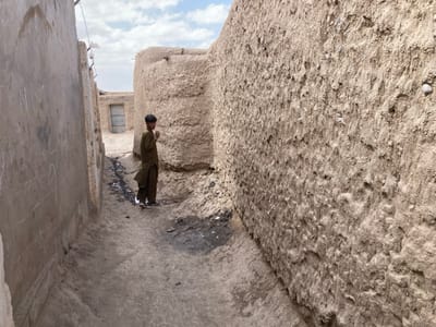 The image shows a narrow alleyway in Saranan, Pakistan, located at coordinates 30.532 latitude and 66.909 longitude. The alleyway is lined by tall, mud-brick walls, one of which has white markings. A young boy in a brown tunic stands at the end of the alley, facing away from the camera. The ground is dirt and there is a small puddle of water in the distance.  The sky is blue and cloudy.