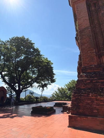 The image captures a bright, sunny day at the Thap Nhan (Nhan Tower) in Tuy Hoa Ward, Vietnam. A section of the ancient red brick tower dominates the right side of the frame, showing weathered textures and vegetation growing in crevices. The foreground features a wide, orange tiled plaza, with several low, rectangular planters, one containing red flowers. Shadows from trees are cast across the plaza.

To the left of the tower, a large, leafy green tree is prominent, its branches reaching towards the sky. Behind the tree, palm trees and other lush vegetation are visible. In the middle ground, a group of three people stand near what appears to be a large, curved red sculpture, with one person wearing a white hat. Beyond the vegetation, a hazy mountain range and a distant town or city are visible under a clear blue sky with a bright sun and lens flare in the upper left corner. The overall atmosphere is tranquil and suggests a popular tourist or historical site.