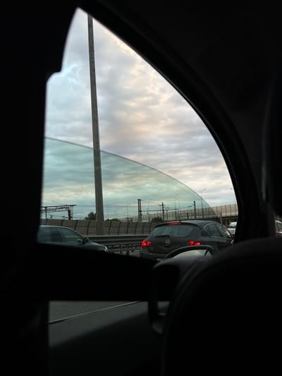 The image shows a view from inside a car, looking out the side window. The scene depicts a traffic jam on a highway, likely in or near Imeni Morozova, Russia, given the license plate of the car in the foreground. The sky is overcast with clouds, and there is a tall pole or tower visible on the left side of the frame. Several cars are stopped on the highway, including a dark gray hatchback with its brake lights illuminated in the center of the image. A portion of the car's interior, including part of the dashboard and seat, is visible in the foreground, obscuring part of the view. The overall atmosphere is one of standstill and muted light, typical of a cloudy day and traffic congestion.