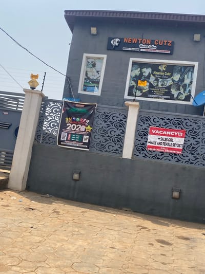 The scene depicts the exterior of a commercial establishment identified as "Newton Cutz Unisex Salon" in Igbessa, Nigeria. The salon occupies the ground floor of a two-story building with a gray stucco facade and a dark brown trim along the roofline.
A black rectangular sign with white lettering and a lion's head logo for "NEWTON CUTZ Unisex Salon" is affixed above the entrance area. Below the main name, smaller text reads "Handsome & Beauty House" and includes Nigerian phone numbers (+2348143410886, +2348033841905). Two rectangular display windows are visible on the first floor. The left window features a graphic of a person getting a haircut. The right window displays a large poster showcasing various hairstyles and listing the salon's services: "UNISEX SALON," "PEDICURE," "FACIAL," and "HAIR TREATMENT," along with additional contact numbers (08147810366, 08033841905). A blue satellite dish is mounted on the building's right side.
In front of the building, a low gray wall with light-colored concrete pillars is topped by an ornate dark gray metal fence featuring a scrollwork design. An electric fence system is installed above the metal fence, with wires and yellow insulators. A yellow, lantern-style light fixture is present on the leftmost pillar. Two smaller, dark-colored wall lights are fixed to the gray wall below the metal fence.
Two banners are affixed to the metal fence. On the left, a black banner advertises a "SNOOKER COMPETITION 2020," detailing prize money (1st - 100K, 2nd - 50K, 3rd - 20K, 4th - 10K, Daily Winner - 5000), and a contact number (09036937684). On the right, a red banner with white capital letters declares "VACANCY!!!" and seeks "SALES GIRL" and "MALE AND FEMALE STYLISTS," providing two contact numbers (08026472628, 09036937684).
The ground in the foreground is paved with reddish-brown interlocking hexagonal pavers. Scattered debris, including plastic and paper, is visible on the pavement. Black electrical wires traverse the upper left portion of the frame. The sky is bright and clear. No individuals are present in the image.