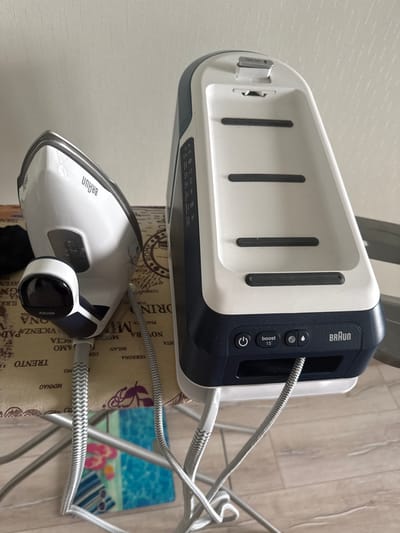 The image shows a Braun steam iron and its base station placed on an ironing board. The steam iron is white with navy blue accents and the Braun logo prominently displayed. The base station is also white and navy blue, featuring buttons labeled "BRAUN", "boost 15", and a power button. A power cord is visible, connecting the iron to the base. The ironing board has a patterned surface with text that appears to list various Italian cities such as Rome, Milan, and Verona, along with other words in Italian. In the background, a blurry image of a beach scene with lounge chairs and flip-flops can be seen on a blue mat. The setting appears to be indoors, likely a home, with a textured wall visible in the upper left corner and a wooden floor beneath the ironing board. The lighting suggests it is daytime. There are no people visible in the image. The overall impression is of household appliances ready for use.