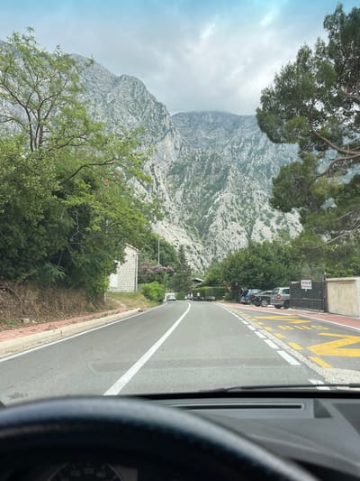 The image displays a scenic mountainous landscape, likely captured from inside a vehicle, with a distinct lean suggesting it was originally a portrait-oriented photograph presented here rotated ninety degrees clockwise.

In the foreground, the dark interior of a car, including part of the dashboard and a pillar, is visible on the far right edge. To the left of this, an asphalt road curves gently into the distance. It features a continuous white line along its inner edge and a dashed white line marking a lane or shoulder. Adjacent to the road, on the right, is a red-paved area with large yellow letters clearly spelling "BUS."

On this red shoulder, several vehicles are parked, including a silver SUV and a dark sedan. Next to the parked cars stands a black metal gate, bearing a white sign with prominent red text that reads "FOR SALE," followed by a partially visible phone number.

The mid-ground is filled with lush green trees and bushes, behind which a small, stone-built house with a white door is nestled. The background is dominated by a majestic, rugged mountain range composed of greyish-white rock, sparsely covered with green vegetation. The upper sections of the mountains are partially obscured by low, grey clouds or mist, indicating an overcast or partly cloudy day. The overall scene evokes a sense of traveling through a picturesque, natural environment in Donji Orahovac, Montenegro.