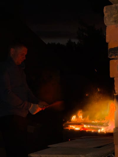 A man stands in front of a bonfire at night in Ol'khovo, Russia. He is wearing a dark jacket and white gloves, and he appears to be holding a tool or stick, possibly tending to the fire. The fire is burning brightly, with flames reaching upwards and embers glowing. The background is dark, with trees and a dark sky visible. A brick structure is present on the right side of the image. The overall scene suggests a domestic or outdoor setting where a fire is being maintained.