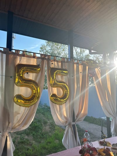 In Pribrezhnyi, Russia, a celebratory outdoor scene unfolds under a covered patio. Two large, golden balloons, shaped like the numbers "5" and "5," are prominently displayed, suspended by string lights. These balloons suggest a 55th birthday or anniversary celebration.

Behind the balloons, light beige curtains hang, framing the view of the outdoors. Through the gap in the curtains, a bright sunbeam cuts across the scene, creating lens flare and casting a warm glow. The background reveals lush green grass and trees, with a glimpse of a light blue sky. A light-colored metal fence is also visible in the distance.

The foreground features a pink tablecloth draped over a table, hinting at a prepared feast. A bouquet of flowers in shades of orange and deep red sits in a dark basket. Various dishes, including fruit and what appears to be charcuterie, are arranged on the table, ready for consumption. A clear plastic water bottle stands near the floral arrangement. The overall impression is one of an outdoor gathering, likely a birthday party, set in a suburban or dacha environment.