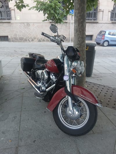 A parked, red and black Harley-Davidson motorcycle is shown on a paved sidewalk in Ávila, Spain. The motorcycle has saddlebags, chrome detailing, and a visible "DELUXE" inscription on its front fender. A light blue car is parked on the street behind the motorcycle, and a trash receptacle stands nearby. A tree trunk with a visible blue accessibility symbol on a pole is situated to the left of the motorcycle. The background features a stone building with architectural details.