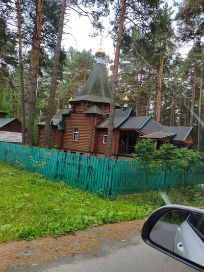 The image shows a wooden Orthodox church with a green roof and golden domes, surrounded by pine trees. A bright turquoise wooden fence runs along the front of the property. The church appears to be made of logs and has traditional architectural elements, including arched windows and a steeply pitched roof. The scene is set in a wooded area, likely a park or a forest, with lush green grass and foliage. A car's side mirror is visible in the foreground, reflecting a portion of the road and another vehicle. A sign is attached to the fence, partially obscured by trees, with Cyrillic text that appears to be related to a church or monastery. The overall impression is one of a serene and somewhat secluded religious site within a natural setting.