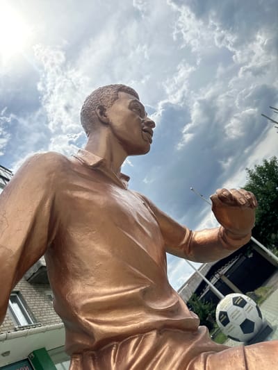 This media file features a bronze statue of a man, angled from a low perspective looking up towards a cloudy sky. The statue depicts a muscular man with his left arm raised in a fist, possibly in a gesture of triumph or power. His head is turned upwards, and his mouth is open, suggesting he might be shouting or celebrating. The statue has a metallic sheen, reflecting the ambient light.

In the background, a soccer ball with black and white panels is visible, suspended or mounted on a pole. Some text is present on the soccer ball, but it is largely illegible due to the image resolution and angle. Behind the statue and soccer ball, there are glimpses of buildings with brick facades and windows, as well as some greenery from trees. A white pole or mast is also visible.

The sky is filled with dramatic, grey and white clouds, indicating a possibly overcast or turbulent weather condition. The sun is partially visible, creating lens flare and highlighting the clouds. The overall impression is one of an outdoor public space, possibly a park or square, dedicated to a sports figure or event, given the presence of the soccer ball. The low-angle shot emphasizes the grandeur of the statue against the vast sky. The time of day appears to be daytime, with the sun's position suggesting it could be morning or afternoon. The location context of Lopaskyne, Ukraine, provides geographical grounding for the scene.