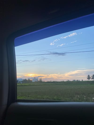 The image shows a view from inside a vehicle, likely a car, looking outwards. The view is segmented by the vehicle's sunroof frame, which is a dark blue color. To the left of the frame, the sky is visible with some scattered clouds and power lines. To the right of the frame, a landscape is visible, featuring a grassy field and distant trees. The setting sun casts a warm glow on the horizon. There is a hint of buildings on the horizon, but their features are indistinguishable. The location is speculated to be near Payarangkuluh, Indonesia.