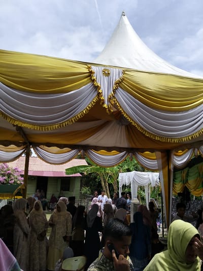 The image depicts an outdoor event, likely a gathering or celebration, taking place under a large white and yellow tent. The tent is adorned with decorative draping and ruffles. In the background, a modest green building with a reddish-brown roof is visible, suggesting a rural or suburban setting in Cottrueng, Indonesia. Several people are present, many of them women wearing headscarves and traditional attire. Some individuals are seen standing, while others appear to be seated or mingling. The overall atmosphere seems to be that of a communal event, possibly a wedding or cultural festival, given the festive decorations and the gathering of people. The sky above is partly cloudy, indicating daytime.