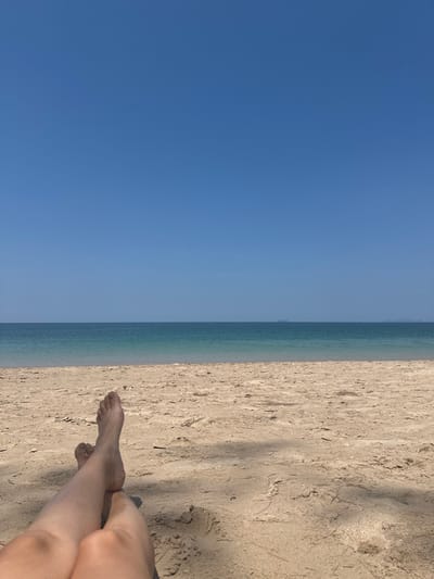 The scene depicts a daytime view from a sandy beach in Phra Ae, Thailand. In the immediate foreground, the bare lower legs and feet of an individual are visible, resting on light-colored sand. The legs are crossed at the ankles, and shadows from an unseen overhead object are cast across the sand. The midground comprises a wide expanse of light brown beach, transitioning into the ocean. The water appears in a gradient, from a lighter turquoise near the shore to a deeper blue further out. The background is dominated by a clear, cloudless bright blue sky, with a distinct horizon line. Two faint, distant landmasses or islands are barely discernible on the horizon against the sky.