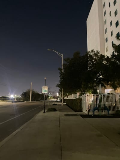 Night scene of a street with a "Speed Limit 30" sign. Street lights illuminate the road, sidewalks, and a building on the left. Trees are visible along the sidewalk and between the building and street. The sky transitions from dark above to lighter closer to the horizon, where distant lights can be seen. A fence and building structure are on the right.
