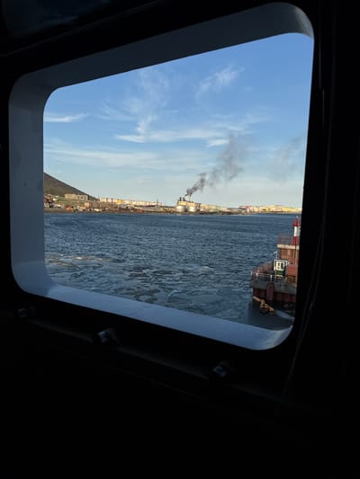 The image, taken from inside a ship's porthole, shows a view of Pevek, Russia. The scene depicts an industrial port with a city skyline in the background. The water is partially covered with ice, suggesting a cold climate. A ship or industrial structure is visible in the foreground to the right, with containers stacked on its deck.
In the distance, several industrial buildings with smokestacks are emitting plumes of dark smoke, indicating ongoing industrial activity. The city extends along the coastline, with numerous multi-story buildings that appear to be residential apartments. The sky is a pale blue with scattered wispy clouds, suggesting it might be late afternoon or early morning.
The overall mood is industrial and somewhat stark, consistent with a port city in a northern region. There is no visible text in the image. The orientation of the image suggests it was taken with the camera rotated.