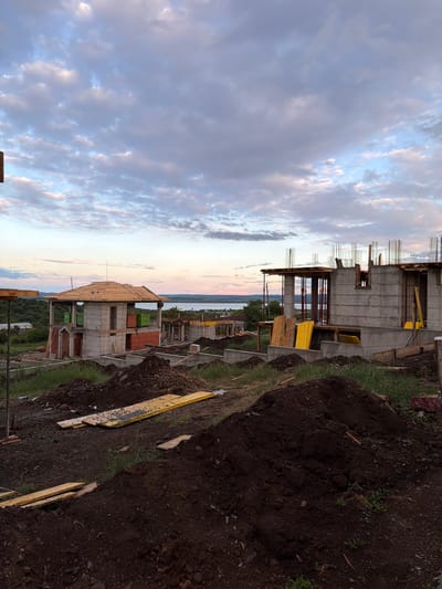 The image shows a construction site in Cherni Vrah, Bulgaria. Multiple buildings in various stages of construction are present, constructed primarily of concrete. Rebar is visible extending from the unfinished concrete structures. The surrounding landscape includes dirt, grass, and construction debris like wooden planks. In the background, there is a glimpse of a body of water and greenery. The sky above is filled with clouds.