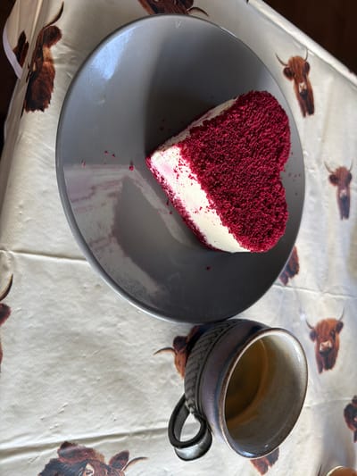 A heart-shaped slice of red velvet cake with white frosting sits on a gray plate. The cake is partially covered in red cake crumbs. A dark-colored mug, with a rough texture and a handle, is placed to the right of the plate. The mug appears to be partially filled with a dark liquid. Both the plate and the mug rest on a tablecloth that features a repeating pattern of Highland cattle illustrations. The overall lighting suggests an indoor setting, possibly a kitchen or dining area. The image does not contain any discernible landmarks or features that would allow for the precise determination of its location within Malvern Hills, United Kingdom.