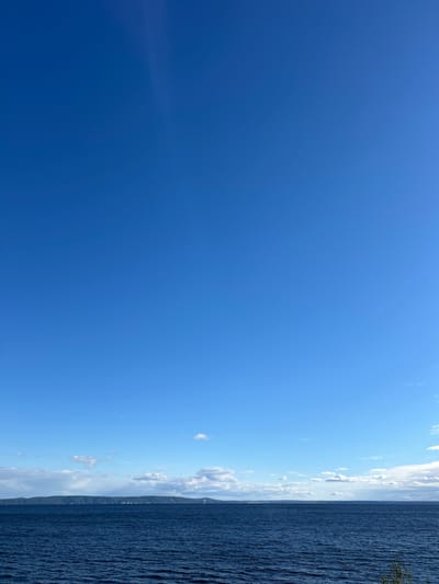 The image depicts a broad expanse of deep blue water, likely a sea or large lake, under a clear, vibrant blue sky. Scattered white clouds dot the upper horizon, and a faint atmospheric haze or contrail is visible in the upper left quadrant. In the distance, a long, low-lying landmass with rolling hills stretches across the horizon, partially obscured by atmospheric conditions. The land appears to be densely forested, with subtle hints of human habitation or structures visible along its shoreline. The overall impression is one of a serene and expansive natural landscape. The image does not contain any people or explicit signs of human activity beyond what might be inferred from the distant landmass. The location is described as being in the city of Primorsky, Russia.
