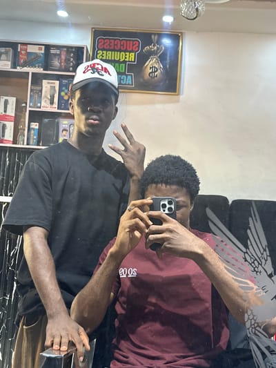 Two men are in a room in Ogwashi-Uku, Nigeria. One man wears a black t-shirt and a white and red baseball cap. The other man is holding up a smartphone and taking the photo. The man taking the photo is wearing a maroon colored shirt. A sign with a dollar sign on it is on the wall. Shelves of hair clippers are also in the background.