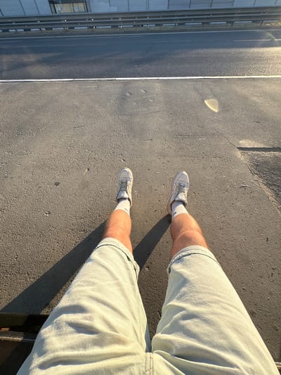 The image is a close-up, overhead shot of a person's legs wearing light-colored denim shorts and white sneakers with white socks. The person is standing on a dark, textured surface, possibly asphalt or concrete. A thin white line runs parallel to the left edge of the frame, separating the textured ground from a darker, smoother surface that appears to be a road or pathway. Sunlight casts long shadows across the ground, suggesting it is either early morning or late afternoon. In the background, to the far left, a building with vertical elements and windows is partially visible, indicating an urban or semi-urban environment. The lighting is bright and warm, illuminating the textures of the clothing and ground, and creating highlights on the road surface. The scene is still, with no apparent activity other than the person standing. There is no visible text in the image. The location context provided is Мирновское сельское поселение, Ukraine.